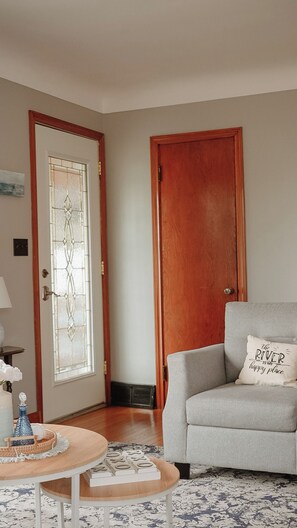 A room with a wooden door, a grey sofa with a pillow, a small round table with a decorative item, and a patterned rug. - The Little River Retreat - Riverfront (St. Clair)