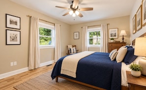 A bedroom with a bed, two windows with curtains, a ceiling fan, and wall-mounted artwork. - Apple Blossom Bungalow - Bike Ride to Ecusta Trail (Hendersonville)