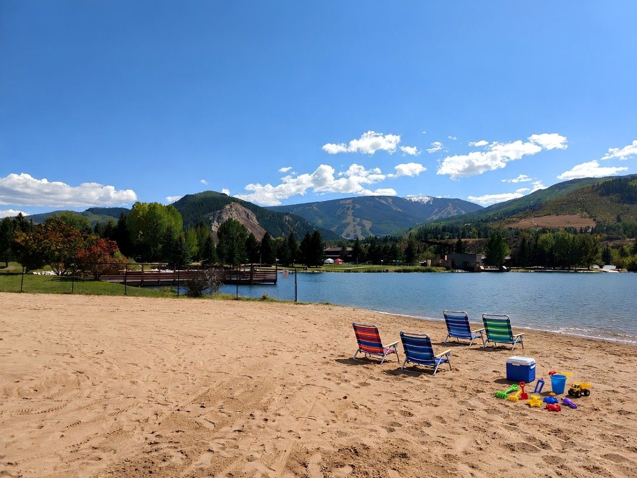 A beach with a lake, mountains in the background, and beach chairs and toys on the sand.