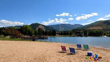 A beach with a lake, mountains in the background, and beach chairs and toys on the sand.