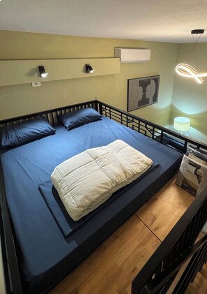 A single bed with a blue mattress and white pillows, a bedside table with a lamp, and a wall-mounted air conditioner. - A comfortable duplex in Fresku, Tirana. (Tiranë)