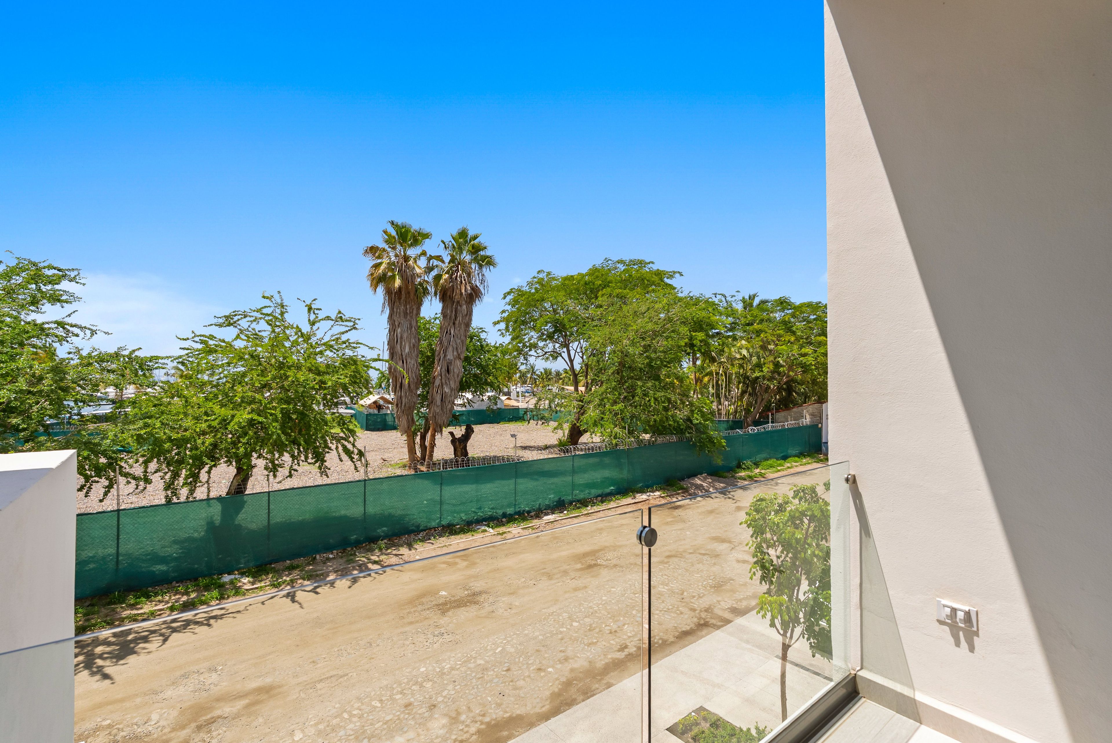 A balcony with a view of a sandy area, palm trees, and a green fence.