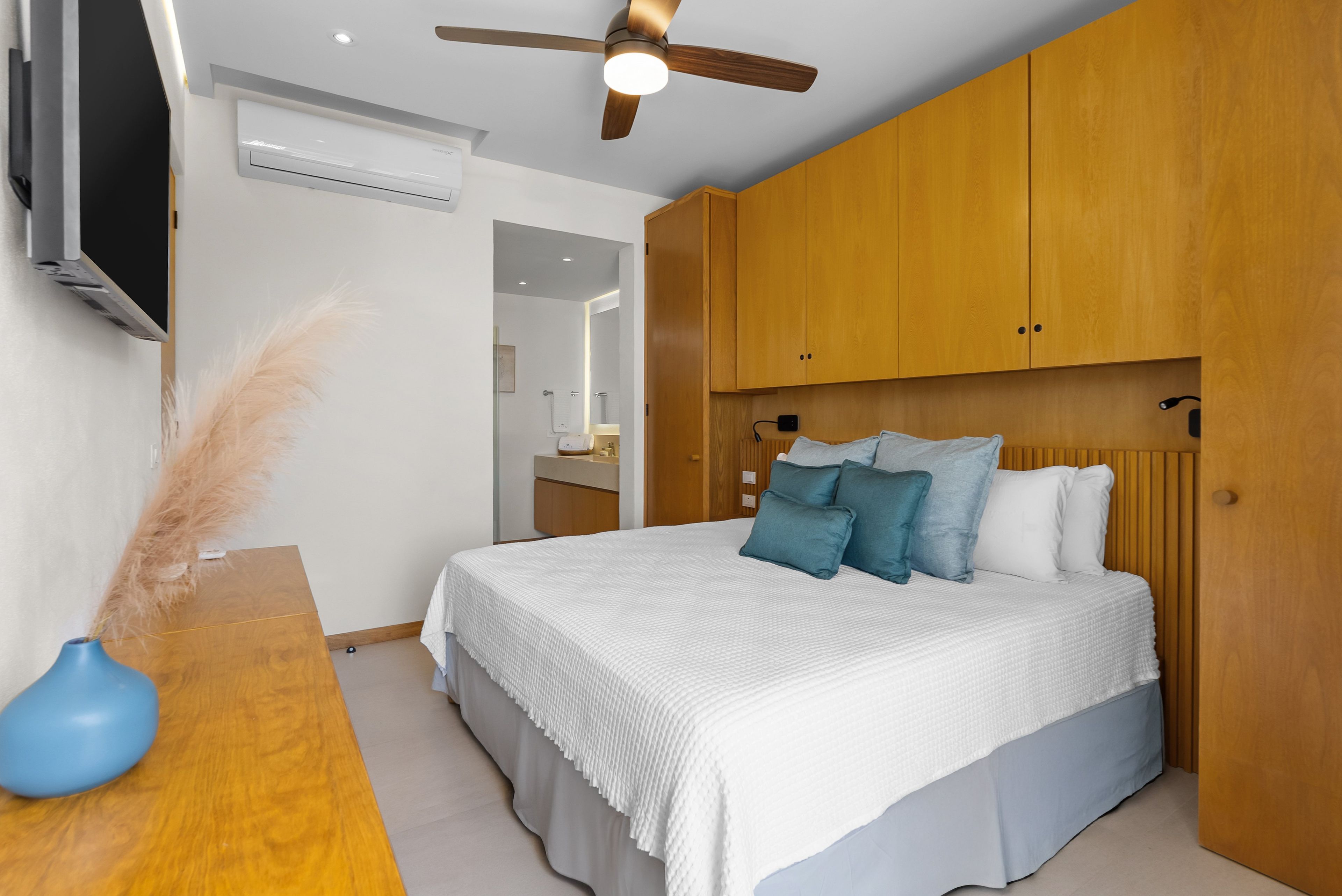 A bedroom with a bed, wooden wardrobe, ceiling fan, and a TV on the wall.