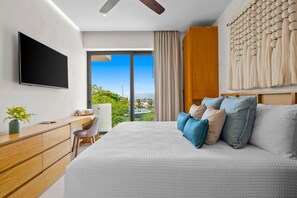 A modern hotel room with a large bed, a desk with a chair, a flat-screen TV, and a view of the outdoors. - 2br, 2BA Alik A5 Condo in La Cruz With Pool, Gym, Balcony, AC, Wifi (La Cruz de Huanacaxtle)
