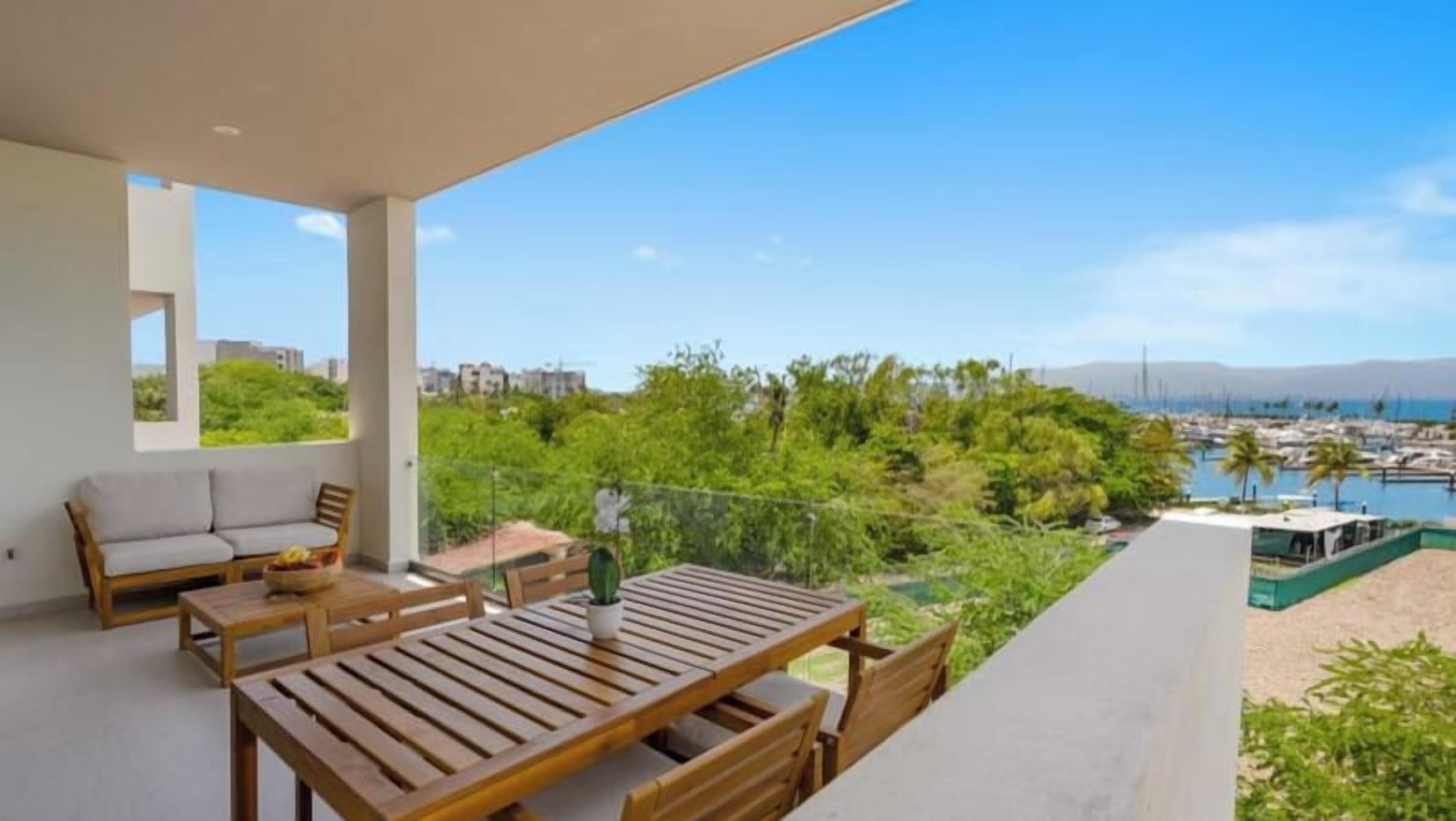 Un balcon avec une table et des chaises en bois, donnant sur une marina et sur un espace verdoyant.