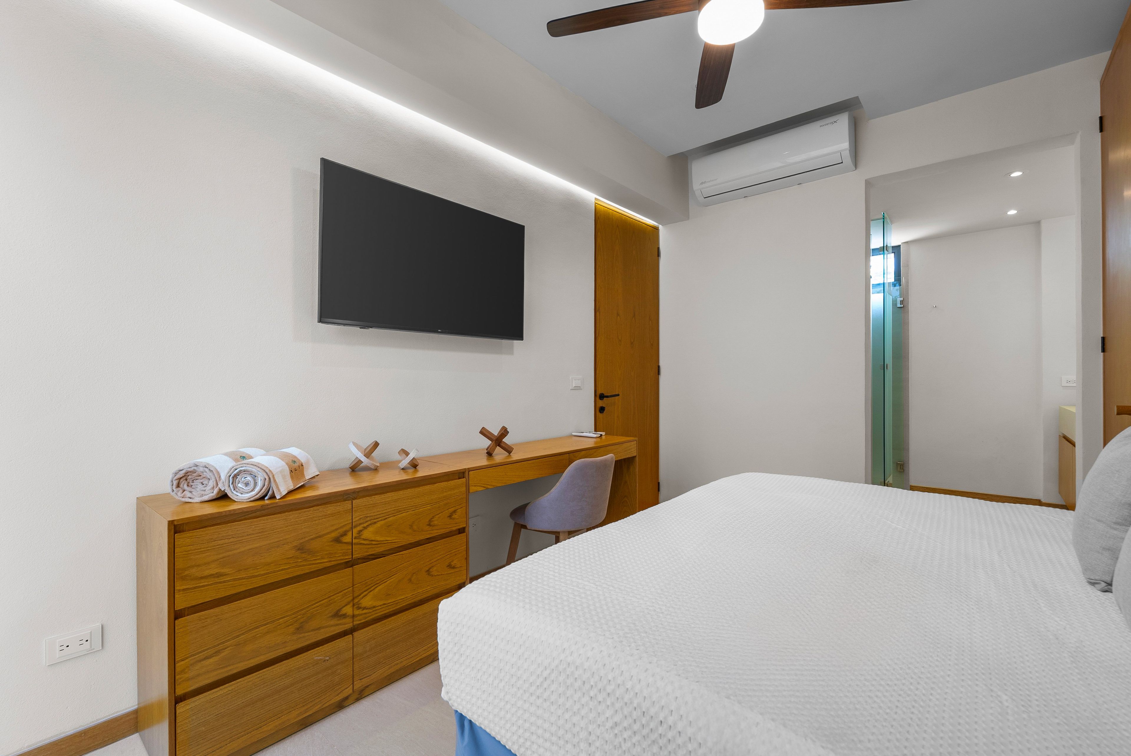 A modern bedroom with a bed, a desk, a chair, a flat-screen TV, and an air conditioning unit.