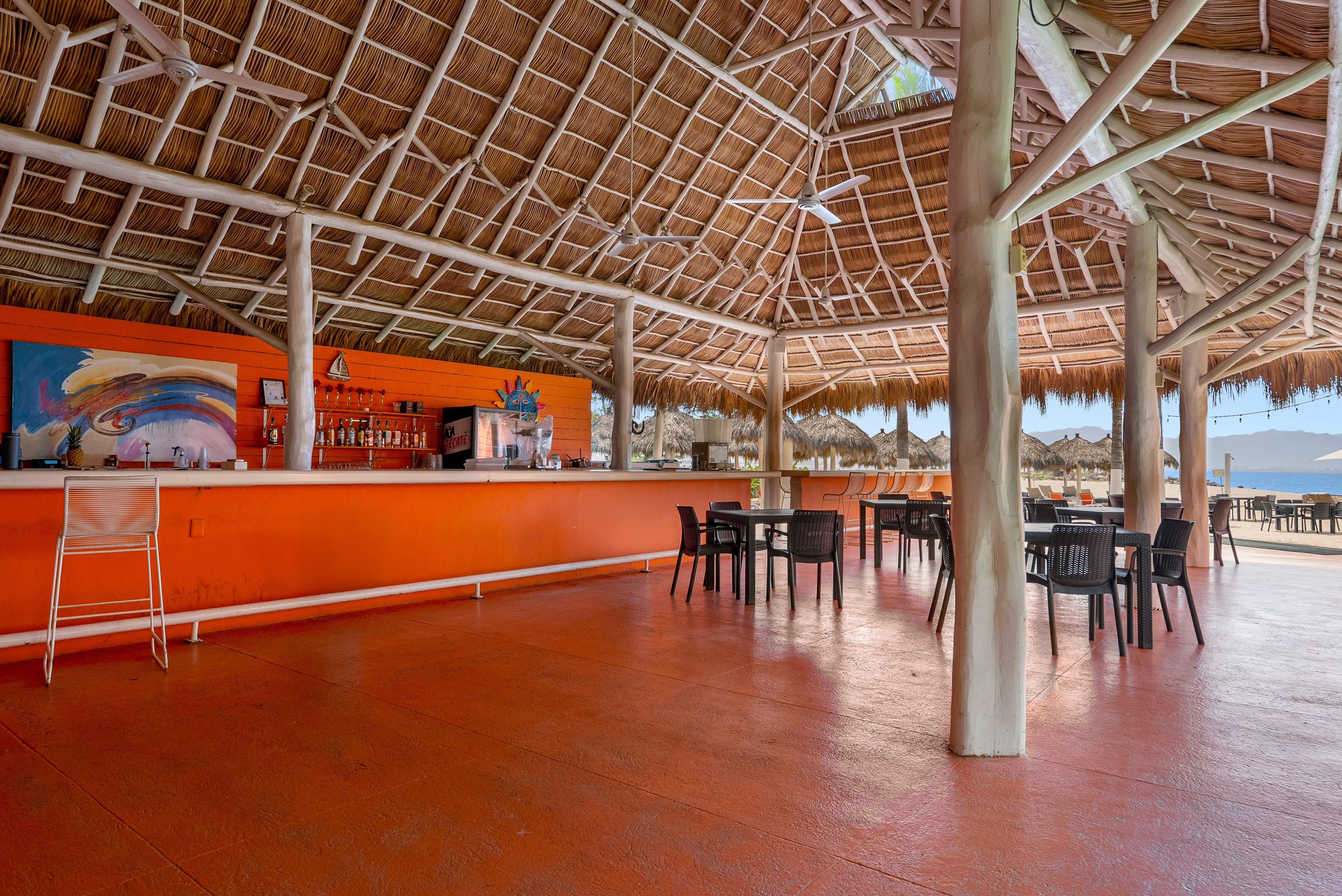 Un área de bar al aire libre con techo de paja, asientos y vista a la playa.