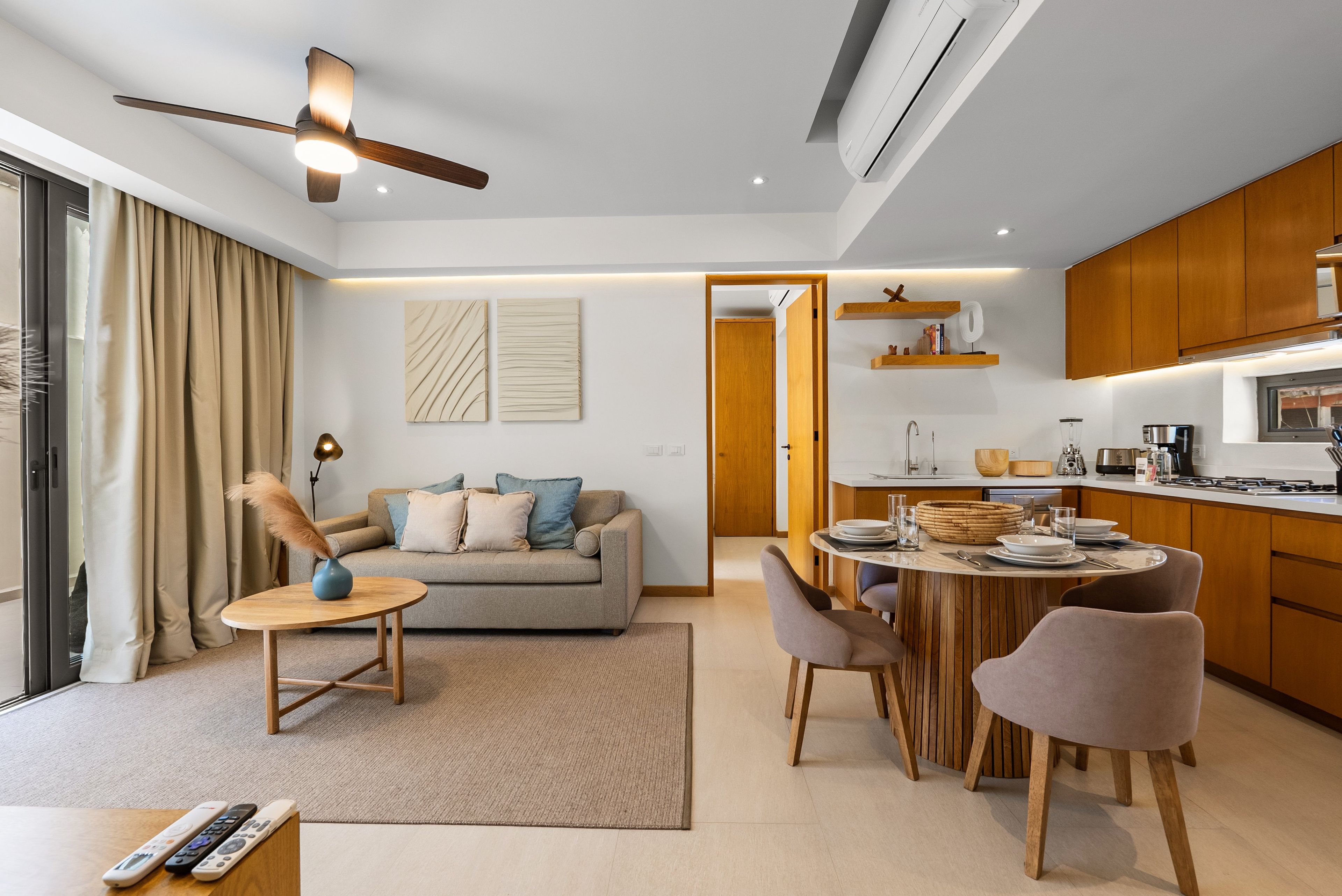 A modern living room with a sofa, coffee table, and dining area with chairs. There is a kitchen with wooden cabinets and a dining table.