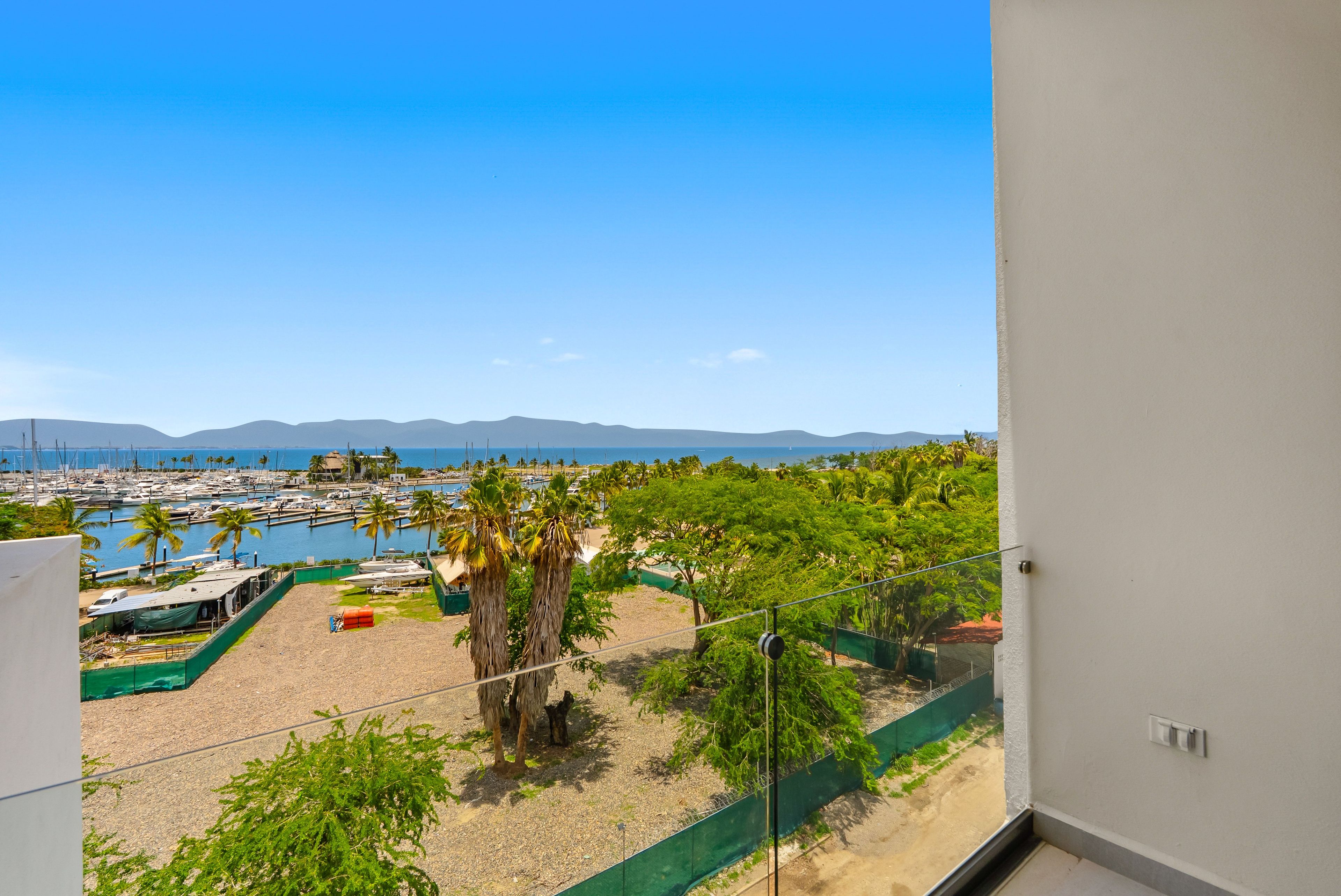 Vista de un puerto deportivo con barcos, palmeras y montañas al fondo.