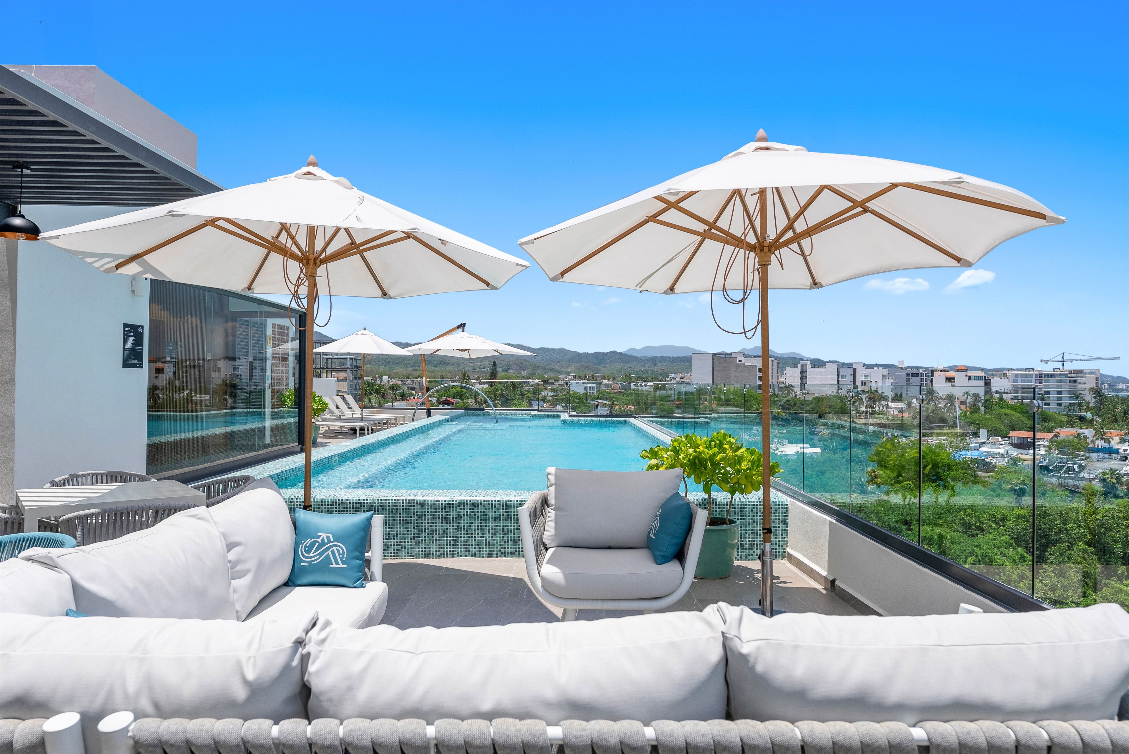 Un área de piscina en la azotea con dos sombrillas, sillones de descanso y vistas a la ciudad.