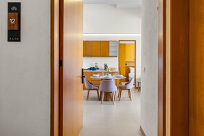 Uma cozinha moderna com armários de madeira, uma área de refeições com mesa redonda e cadeiras, e um corredor que dá para um quarto identificado com o número 12.