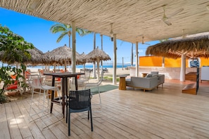 A covered outdoor seating area with tables and chairs, a sofa, and a view of the beach and ocean.