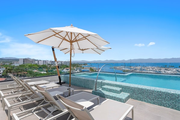 A rooftop pool area with lounge chairs and an umbrella, overlooking a marina and distant mountains.