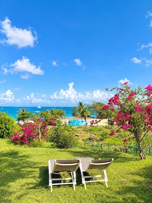 A grassy area with two white lounge chairs facing a pool and ocean view. - Spacious apartment with a garden just a stone's throw from the beach (Saint Martin)