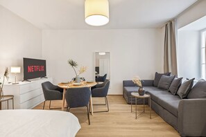 A modern hotel room with a bed, a sofa, a dining table with chairs, a TV, and a window with curtains. - Apartment with a view of the garden & air conditioning | Danube Canal | 6 guests (Wien)