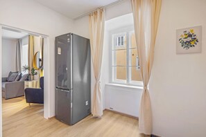 Une cuisine moderne avec un réfrigérateur en acier inoxydable, un coin repas doté d’un canapé gris et une fenêtre avec des rideaux.