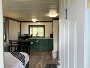 A compact room with a bed, a small table, a kitchenette with a stove, and a window. - Your Cozy Studio with Open Scenic Views & Pine Surroundings (Bayfield)