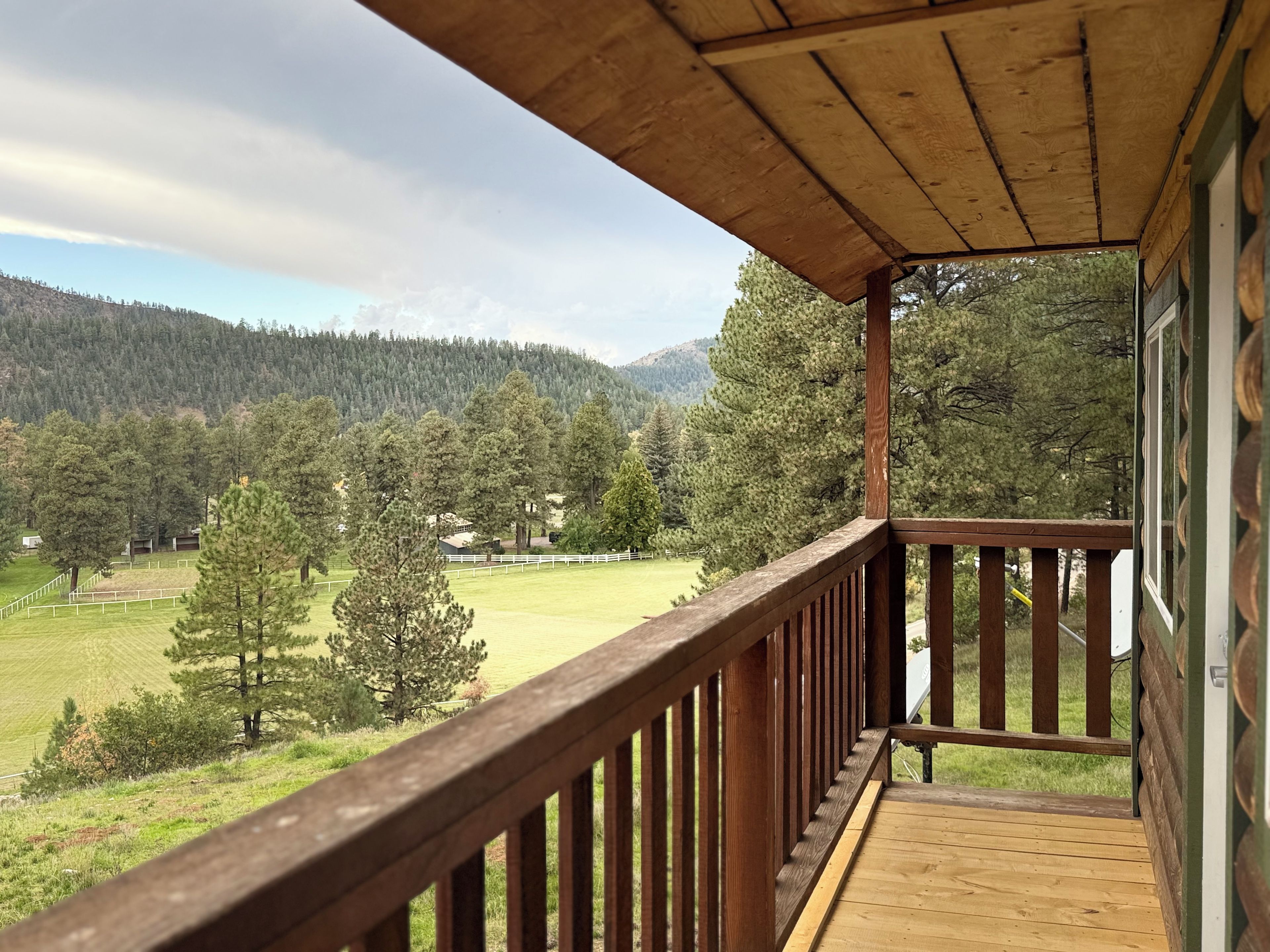 Un balcon en bois offrant une vue sur un champ verdoyant et des arbres, surplombant un paysage vallonné.