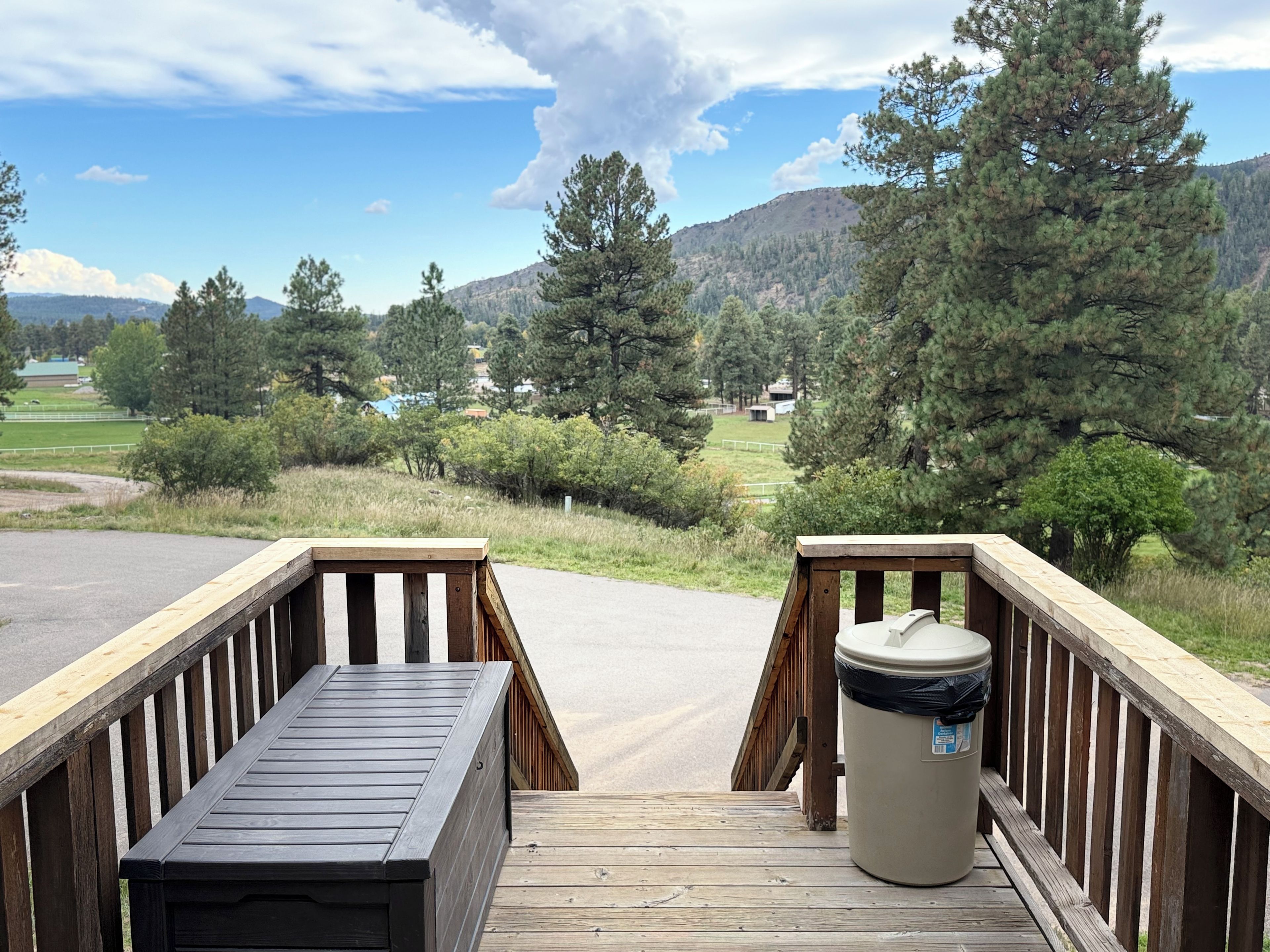 A wooden deck with a trash bin and a bench, overlooking a mountainous landscape with trees and a distant view of a lake.