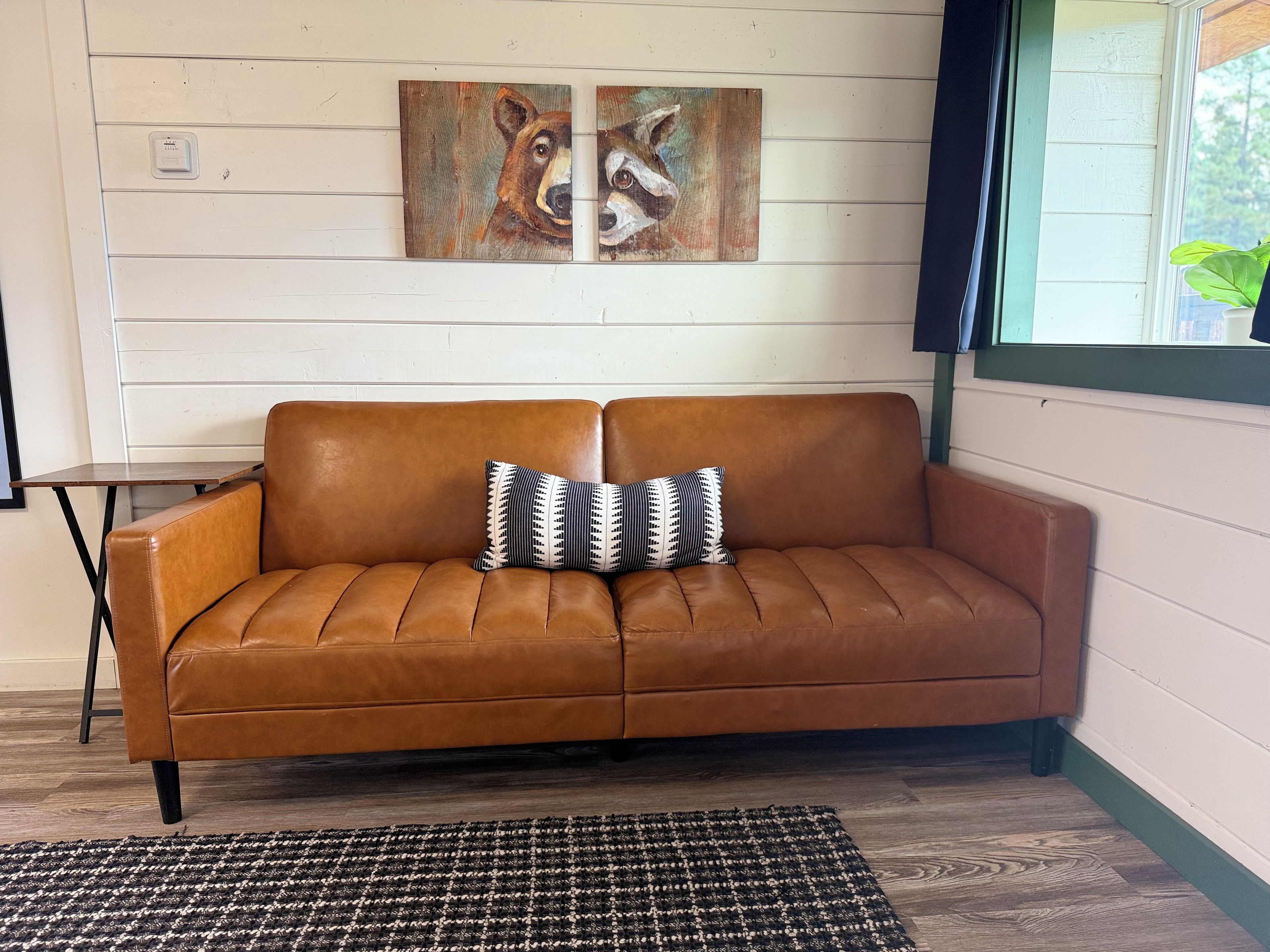 A brown leather sofa with two patterned pillows, a black and white woven rug, and two paintings on the wall.