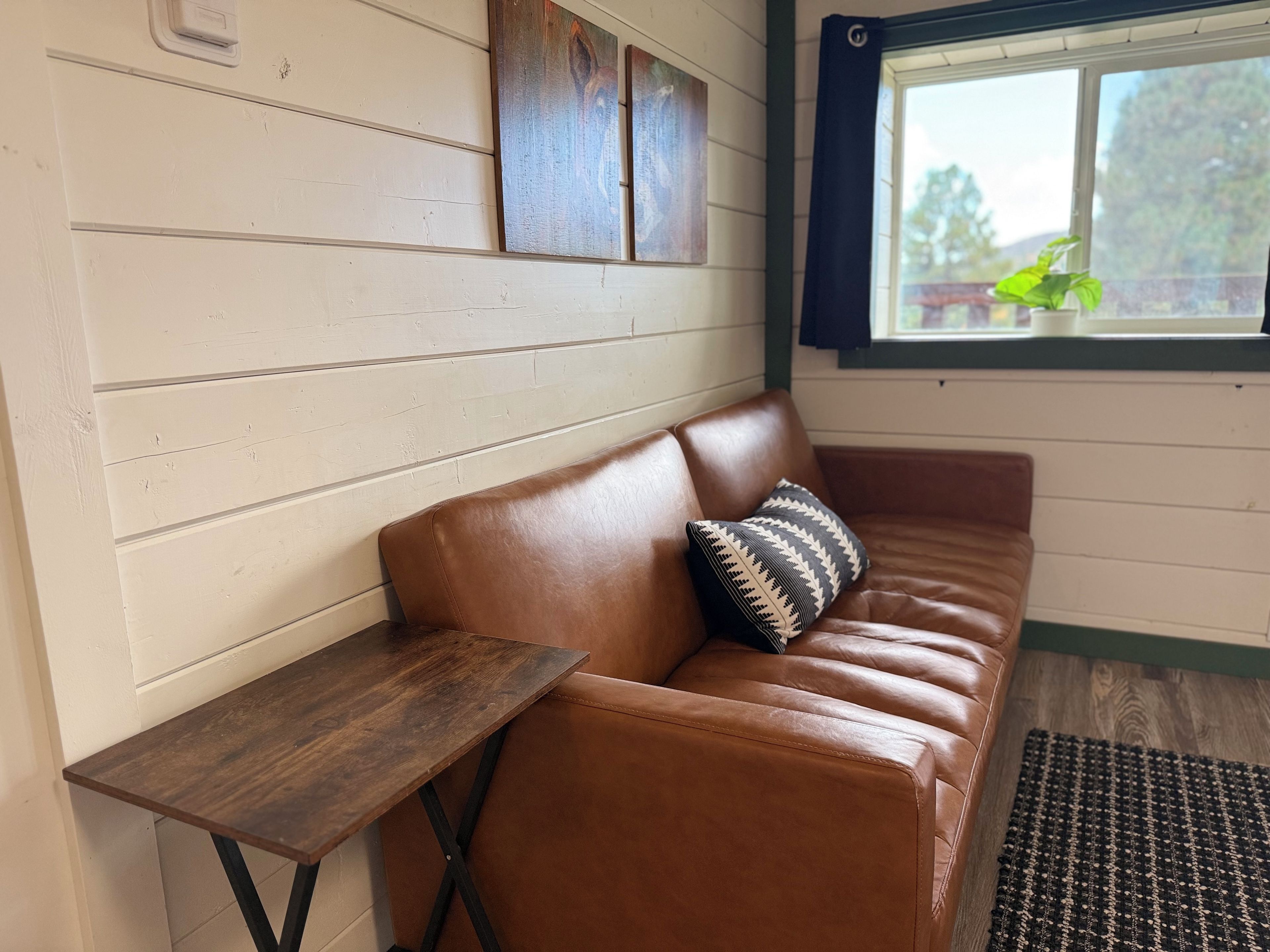 Un canapé en cuir marron agrémenté d’un coussin à motifs, une table d’appoint en bois et une fenêtre donnant sur un paysage verdoyant.