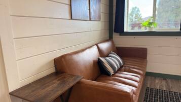 A brown leather sofa with a patterned pillow, a wooden side table, and a window with a view of greenery.