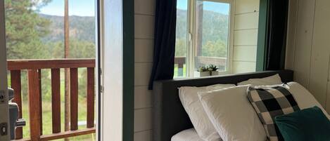 A bedroom with a bed, a window with curtains, a view of the outdoors, and a wooden deck outside.