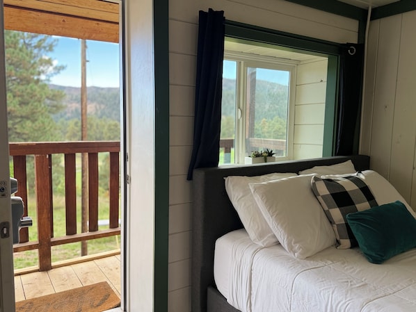 A bedroom with a bed, a window with curtains, a view of the outdoors, and a wooden deck outside.