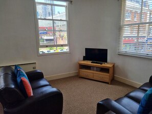 Ein Zimmer mit Fernseher auf einem Holzboden, zwei schwarzen Ledersofas, einem Fenster mit Jalousien und einem Teppichboden.