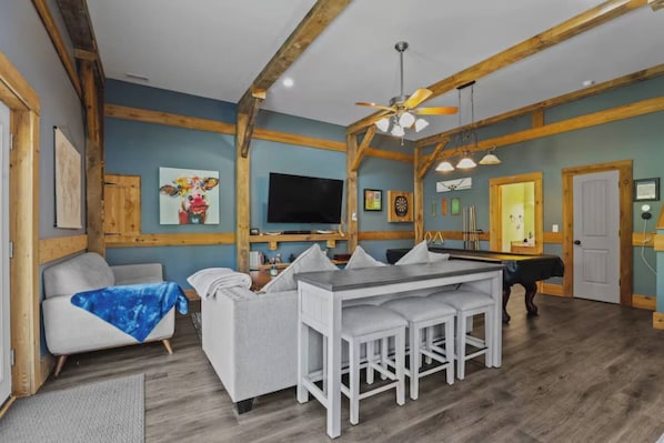 A room with a sofa, a dining table with chairs, a pool table, a ceiling fan, and a television.
