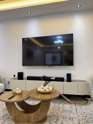 A modern living room with a flat-screen TV mounted on the wall, a wooden coffee table with decorative items, and a white entertainment unit with speakers. - Warn and Cozy 2-bedroom apartment in perfect Dakar-Almadies (Dakar)