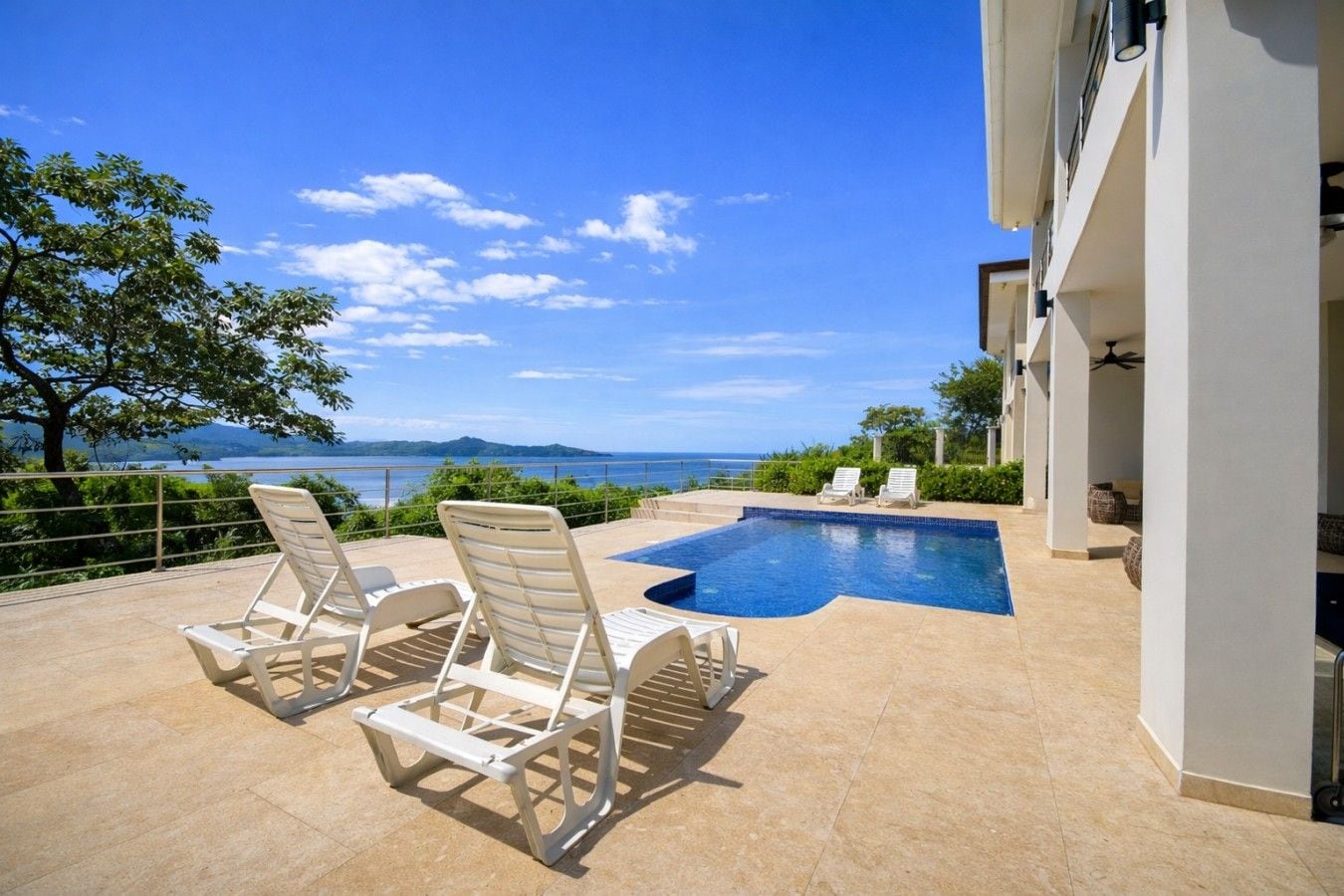 A pool area with lounge chairs, a clear view of the sea, and a building with a balcony.