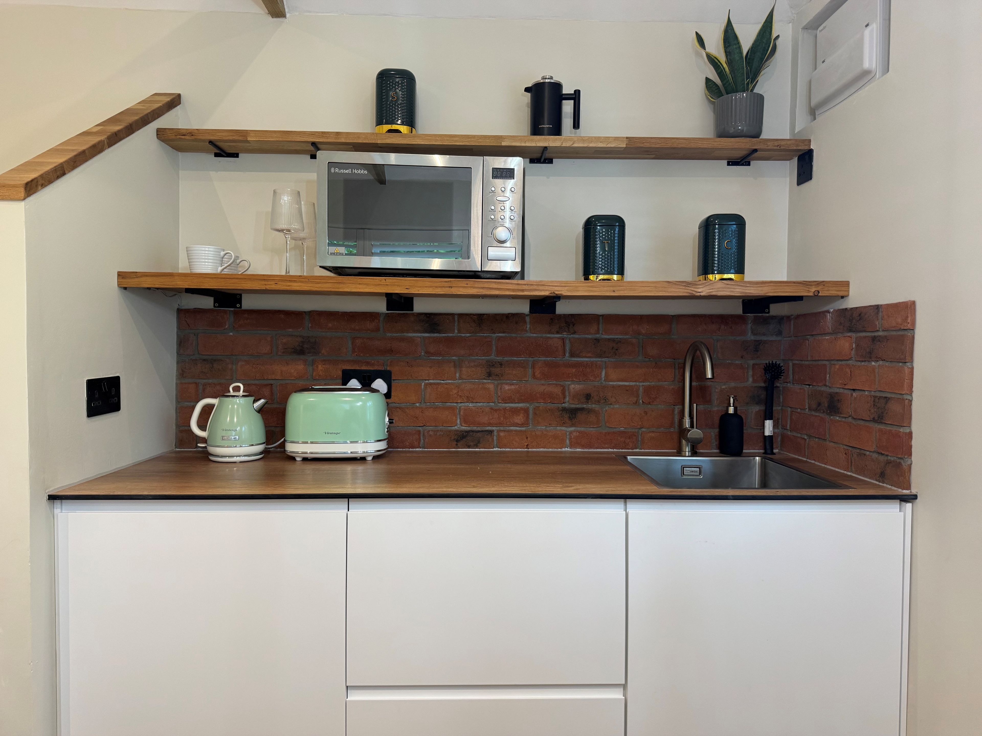 Une cuisine avec un mur recouvert de briques, un four à micro-ondes, un grille-pain, une bouilloire, un évier et une étagère ornée d’une plante et d’une machine à café.
