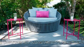 Un sofá redondo de mimbre con cojín blanco y almohada rosa, flanqueado por dos mesitas metálicas rojas con candelabros de cristal, sobre un patio de hormigón rodeado de follaje.
