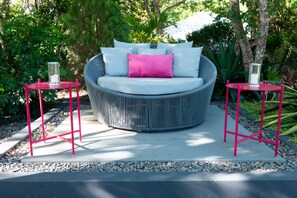 Un sofá redondo de mimbre con cojín blanco y almohada rosa, flanqueado por dos mesitas metálicas rojas con candelabros de cristal, sobre un patio de hormigón rodeado de follaje.