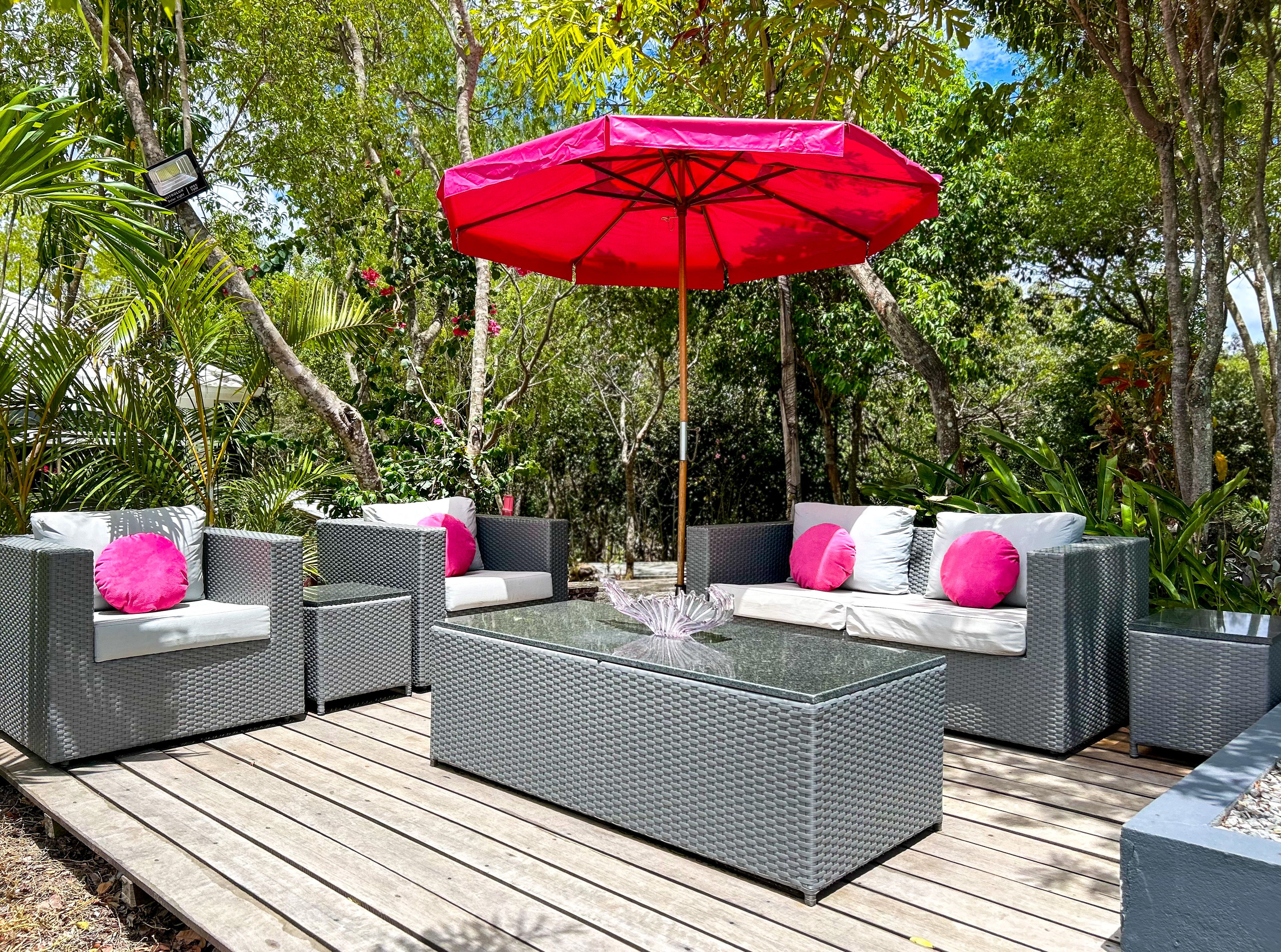 Un patio al aire libre con mobiliario de mimbre, una mesa de vidrio y un paraguas rojo.