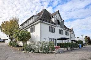 A white building with a steeply pitched roof, multiple windows with shutters, and a sign indicating the name and number. - Morning Light | seeglueck (Reichenau)