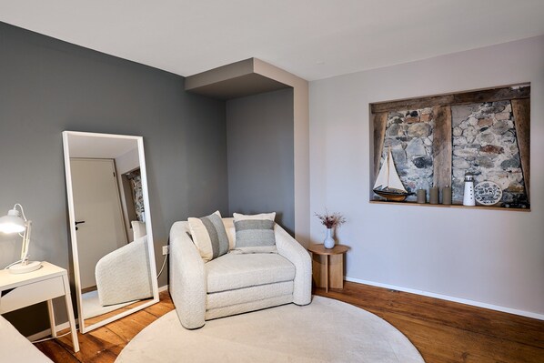 A modern bedroom with a gray sofa, a round rug, a mirror, a small side table with a vase, and a wall-mounted artwork. - Morning Light | seeglueck (Reichenau)