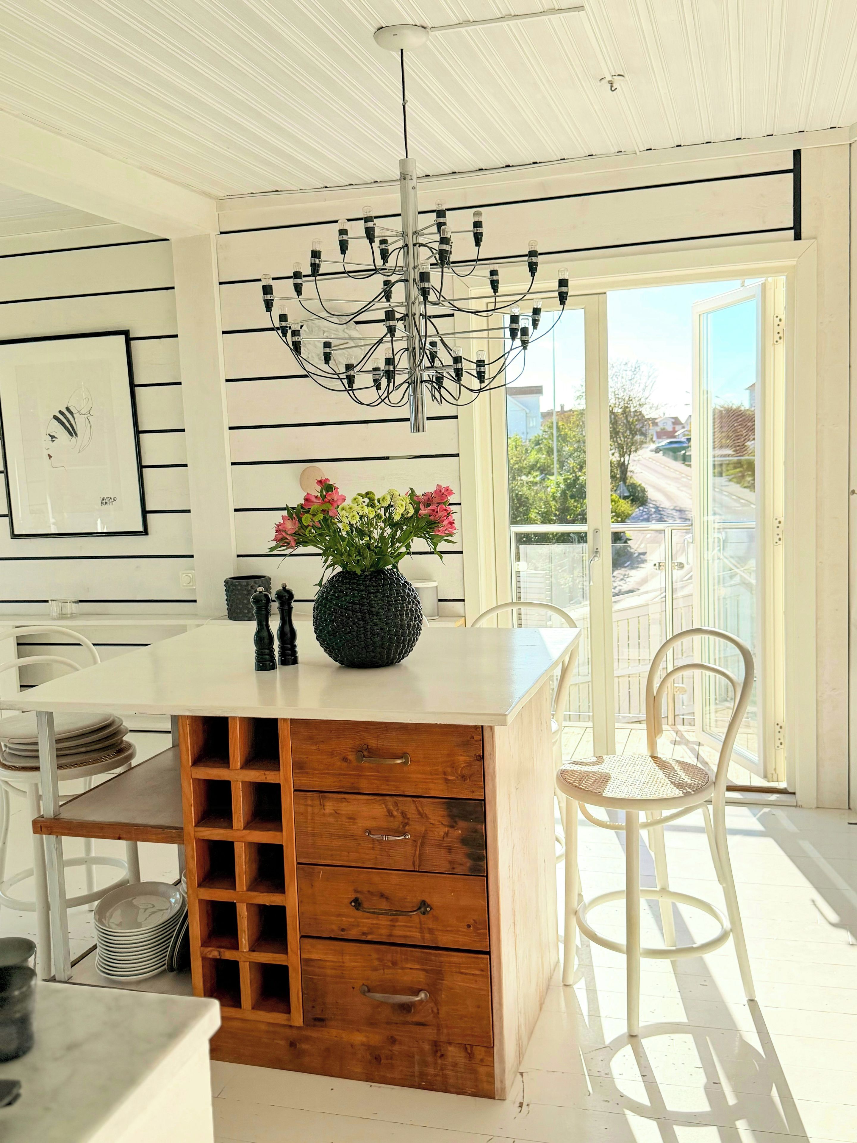 Een keuken met een houten bar, een kroonluchter, een vaas met bloemen en uitzicht door een glazen deur.