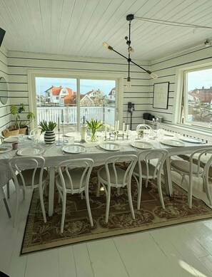 A dining area with a table set for six, white chairs, a patterned rug, and a view of houses through large windows. - House with jacuzzi and sauna on Smögen (Smögen)