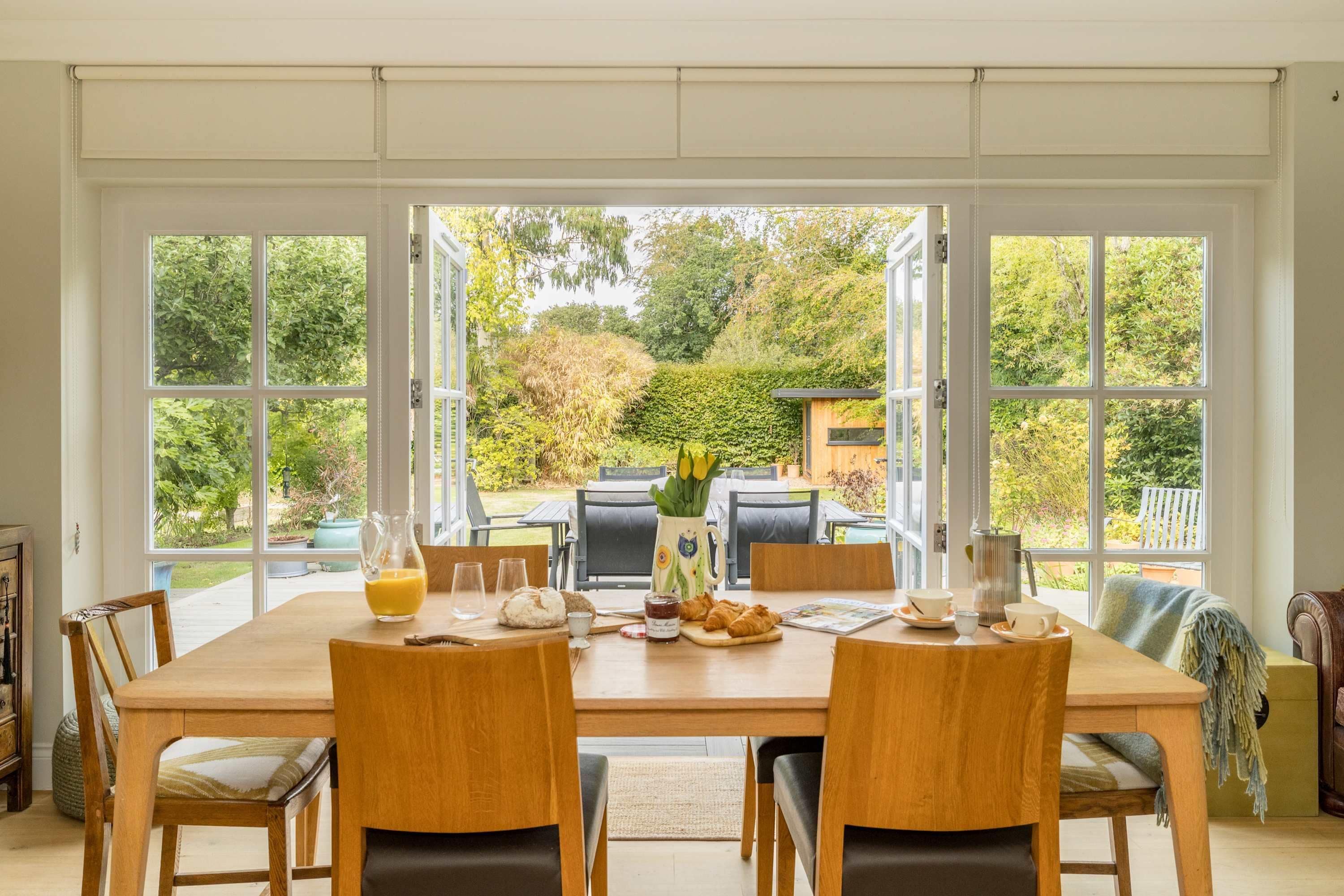 A dining area with a large table, chairs, and a view of the garden through open doors.
