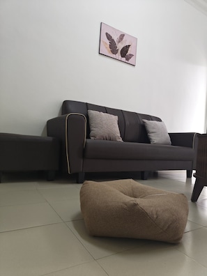 Una sala de estar con un sofá de color oscuro, dos cojines, un taburete tejido y una obra de arte colgada en la pared.