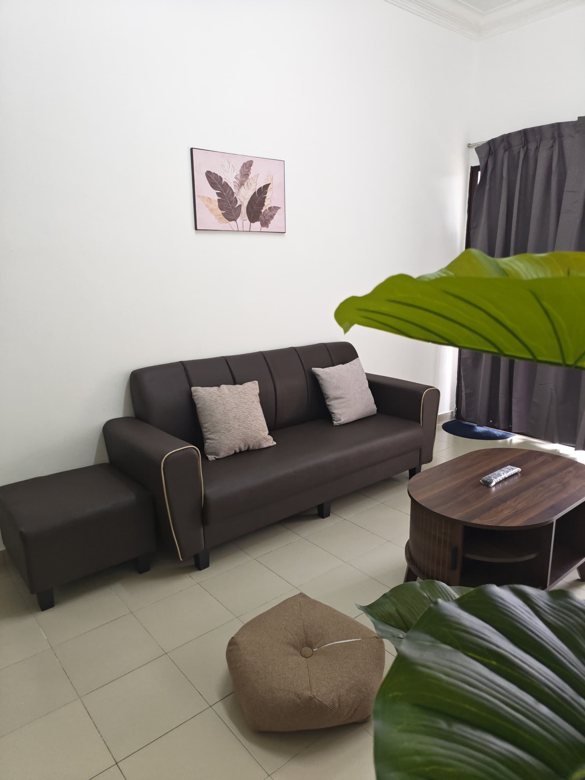 Una sala de estar moderna con un sofá marrón oscuro, una mesa de centro redonda de madera, un otoman beige y una planta verde grande.