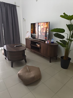Una sala de estar con televisor de pantalla plana, mueble de madera oscura para equipo multimedia, una mesa de centro redonda, un sillón tipo bean bag, una planta en maceta y una cortina.