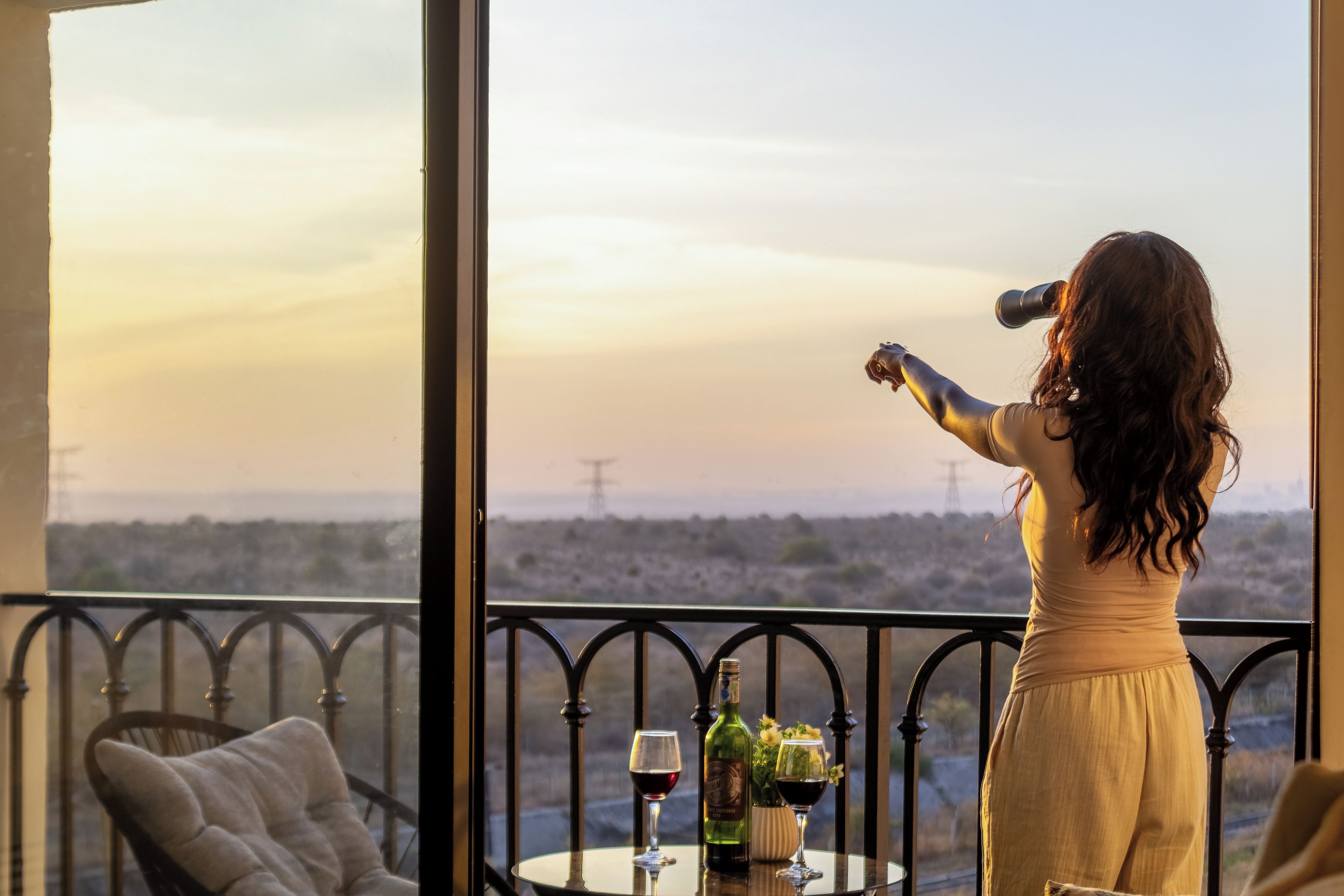 Una mujer de pie junto a un balcón con vistas a un paisaje y tendido eléctrico, una mesa con una botella y una copa, y una silla con cojín.