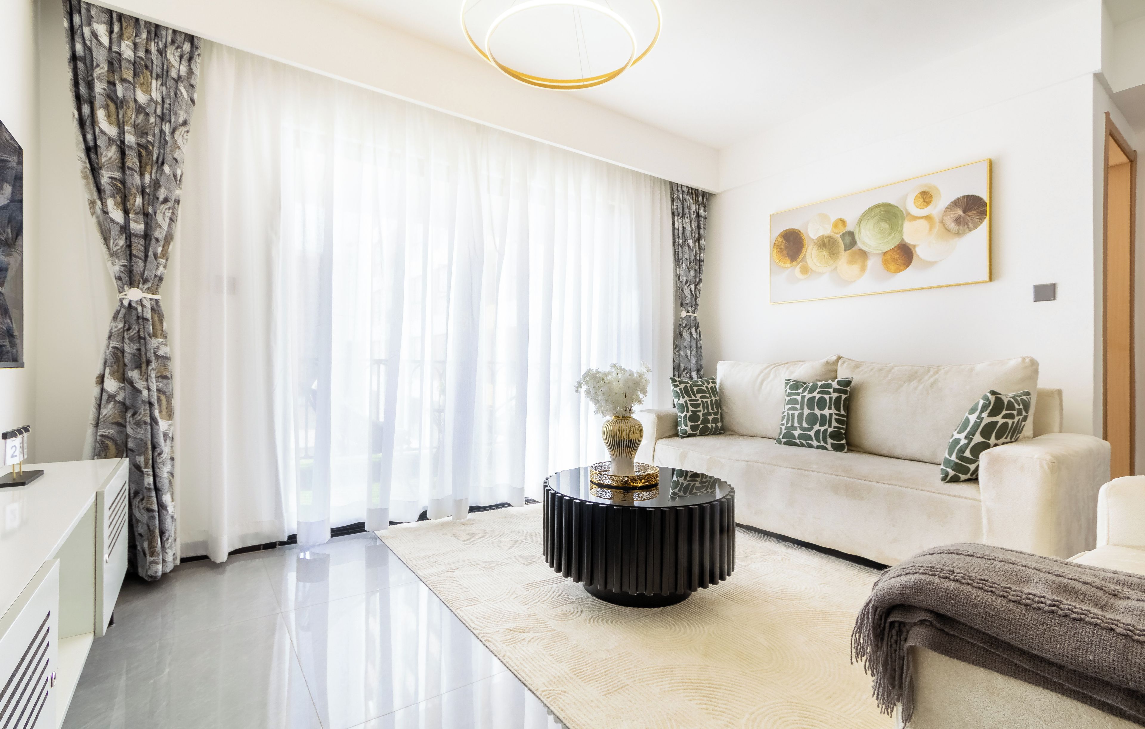 A modern living room with a sofa, a round black coffee table, and a large window with sheer curtains.