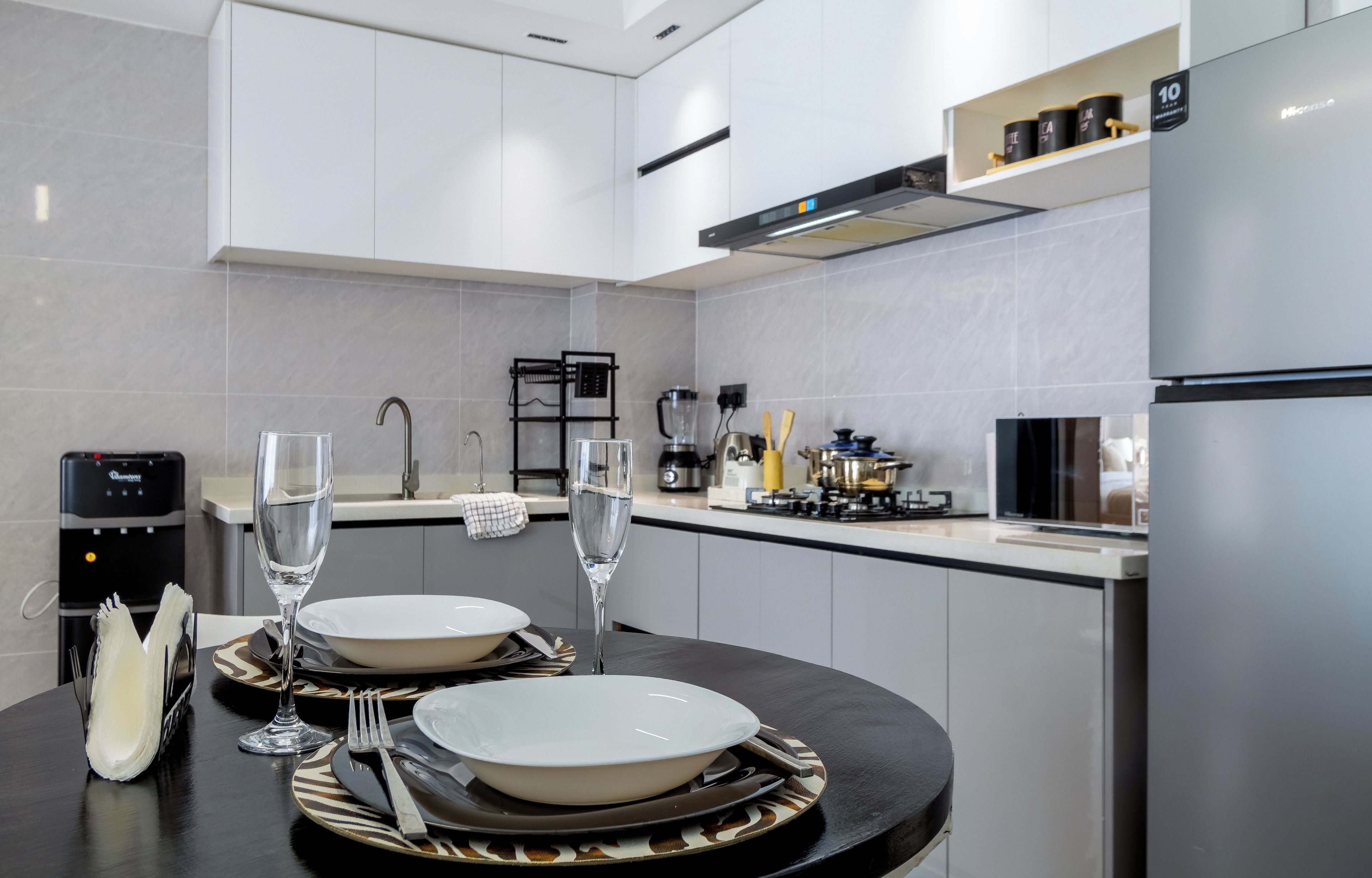 A modern kitchen with a dining area set for two, featuring a water dispenser, a coffee machine, and a built-in oven.