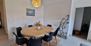 A dining area with a round wooden table, black chairs, a vase with dried plants, a hanging light fixture, and a wall-mounted plant shelf. - Le Cocon Océan – 3-Bedroom Vacation Rental with a Terrace in Moliets (Moliets-et-Maa)