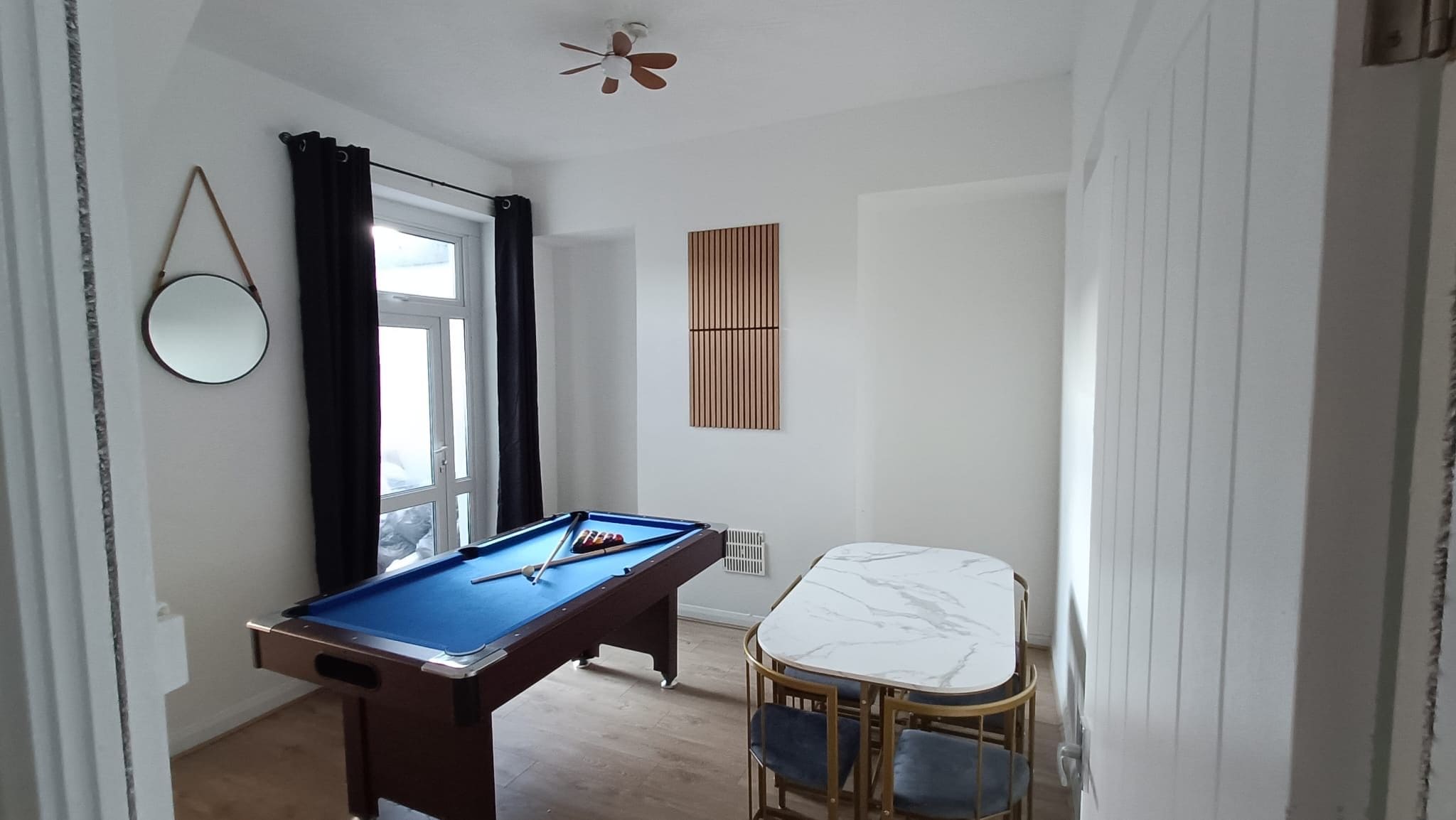 Una habitación con mesa de billar, una mesa de comedor con sillas, un ventanal con cortinas y un ventilador de techo.