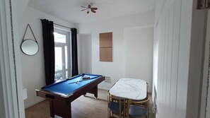 Una habitació amb billar, una taula de menjador amb cadires, una finestra amb cortines i un ventilador de sostre.